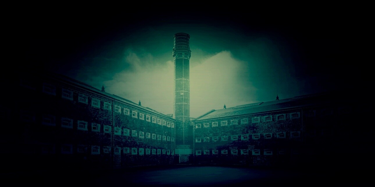 A green-blue tinted image of a tall, external brick tower overlooking three-storey Y-shaped brick prison wings containing dozens of small windows